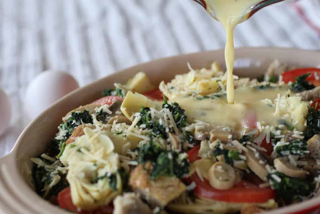 Layers of bread and vegetables in pan with custard being poured over 