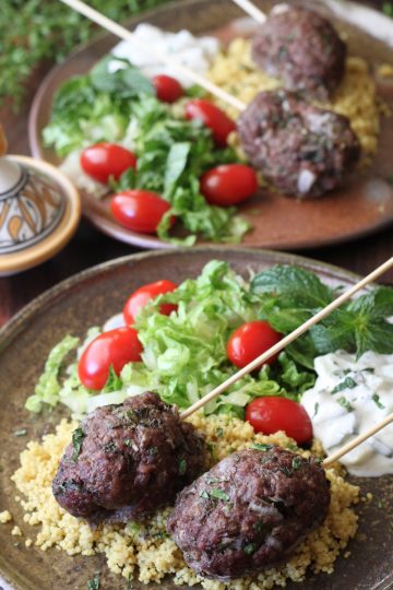 2 plates with ground beef kabobs with lettuce, tomato and couscous and cucumber yogurt sauce.